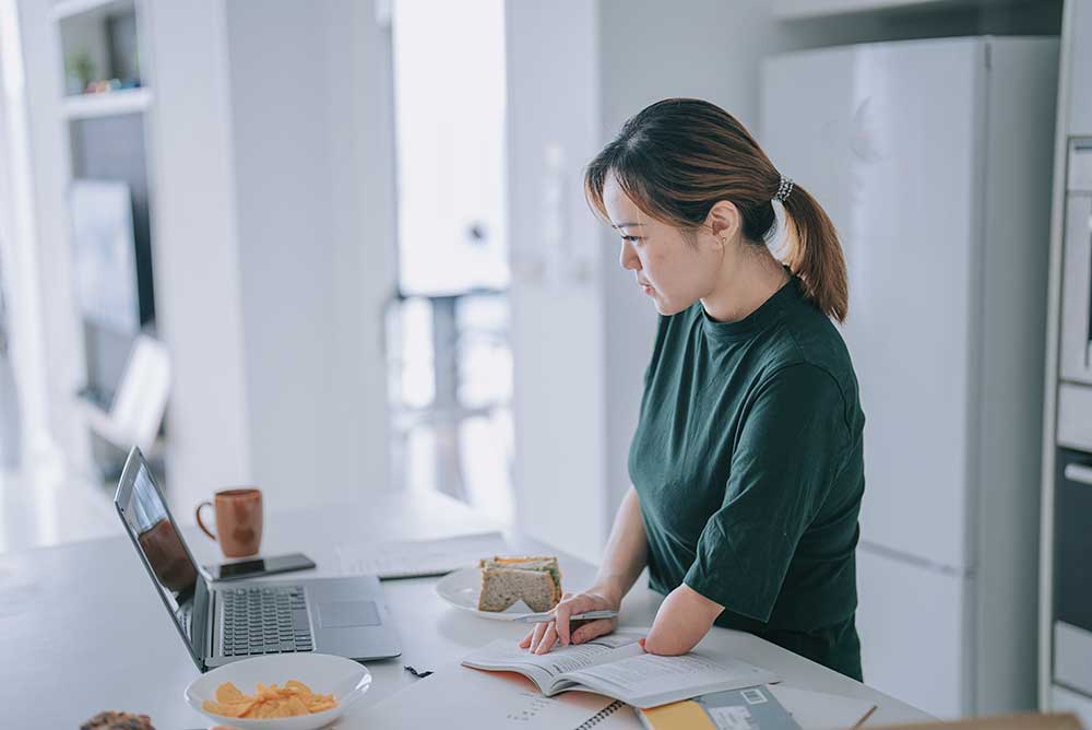 Woman with an arm disability e-learning at kitchen counter using laptop while eating sandwiches