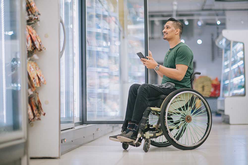 Man with disability on wheelchair shopping at refrigerated section supermarket for frozen food during weekend, checking grocery list on his phone