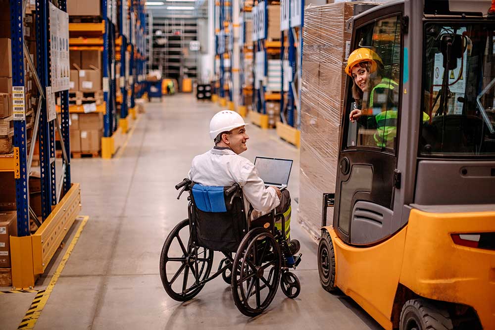 Big industry pallet warehouse with two people working using forklift and laptops and on of them in wheelchair